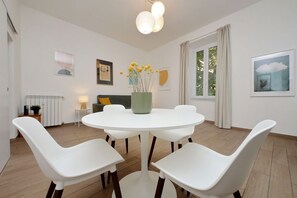 Un comedor con una mesa redonda, cuatro sillas blancas, un florero verde con flores amarillas y una ventana con cortinas.
