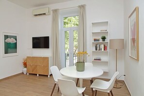 Une pièce moderne avec une table ronde blanche, des chaises blanches, une bibliothèque, un téléviseur et une fenêtre avec des rideaux.