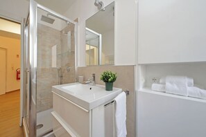 Un baño moderno con cabina de ducha de vidrio, un lavamanos blanco, un espejo y una estantería con toallas.