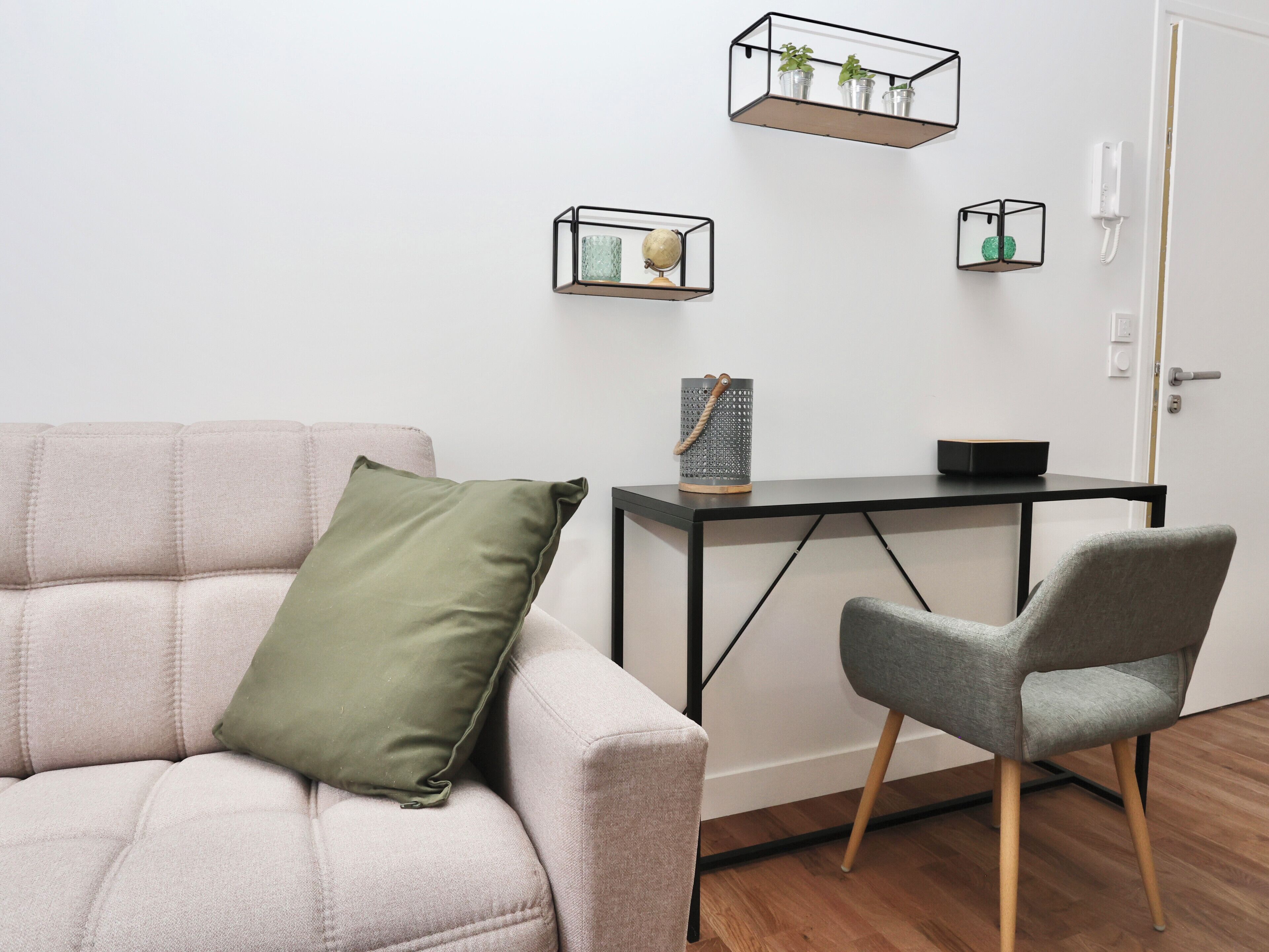 Ein modernes Wohnzimmer mit einem hellen Sofa, einem grauen Sessel, einem kleinen schwarzen Tisch und zwei Wandregalen mit Dekorationselementen.