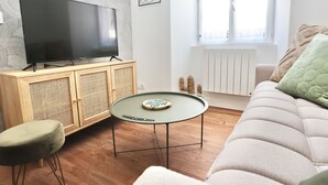A living room with a flat-screen TV on a wooden cabinet, a round coffee table, a green cushioned stool, and a beige sofa with a green pillow.