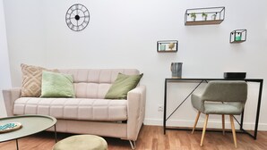 A modern living room with a beige sofa, green cushions, a round ottoman, a small round table, a grey chair, and a black-framed desk with shelves.