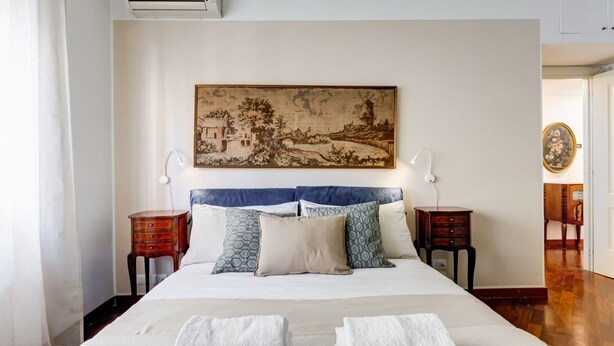 A neatly made bed with white linens, two bedside tables with lamps, a wall-mounted air conditioner, and a framed landscape painting above the bed. - L&L - Central charming 2-Bdr! (Roma)