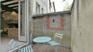 Un patio al aire libre con una mesa redonda y dos sillas, una pared de ladrillo con una pequeña ventana y una puerta que da a un espacio interior con mostrador de cocina y sillas.