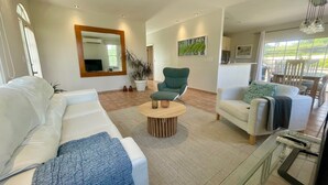 A modern living room with a white sofa, a green armchair, and a glass coffee table. There is a large mirror on the wall, a potted plant, and a painting. - Tierra del Sol Beach Home (Noord)