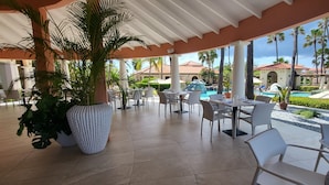 A covered outdoor seating area with tables and chairs, surrounded by potted plants and overlooking a pool and palm trees. - Tierra del Sol Beach Home (Noord)