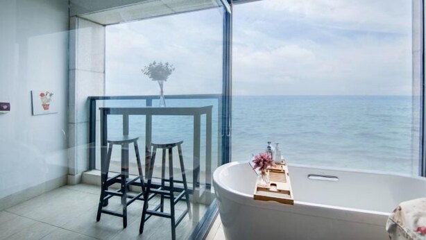 A modern bathroom with a large bathtub, a glass-enclosed balcony with a view of the sea, and two stools.