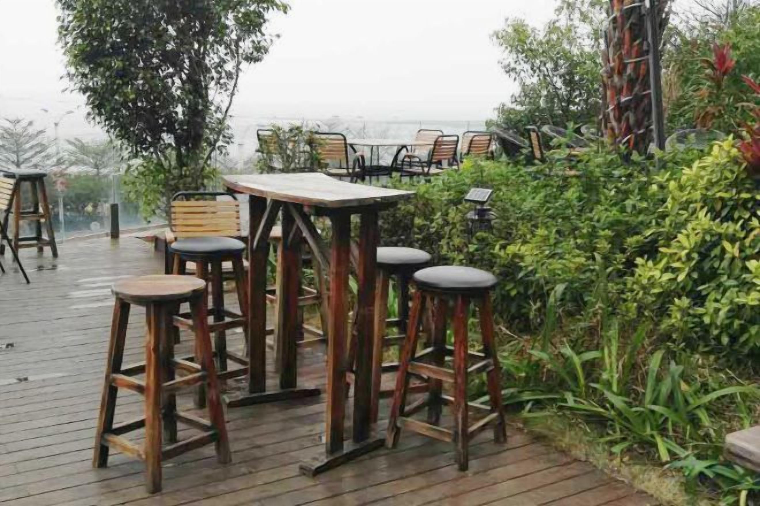 An outdoor seating area with wooden tables and stools, surrounded by greenery and overlooking a view.