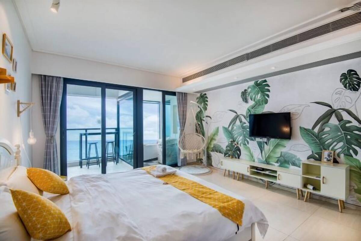 Una habitación de hotel moderna con una cama grande, televisor, balcón con vistas y un mural temático tropical.