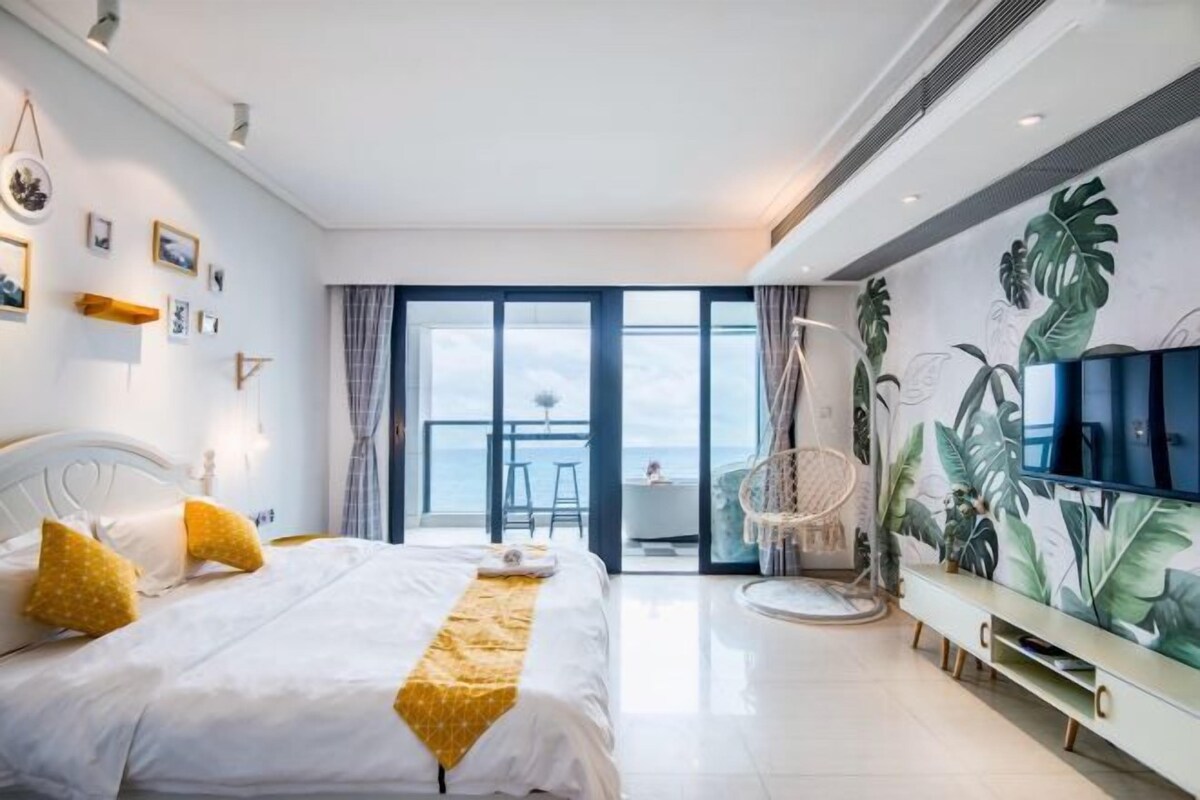 A modern hotel room with a large bed, a TV, a balcony with a view, and a tropical-themed wall.