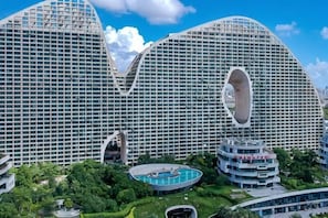 A modern hotel with a distinctive curved facade, a central swimming pool, and lush surrounding greenery.