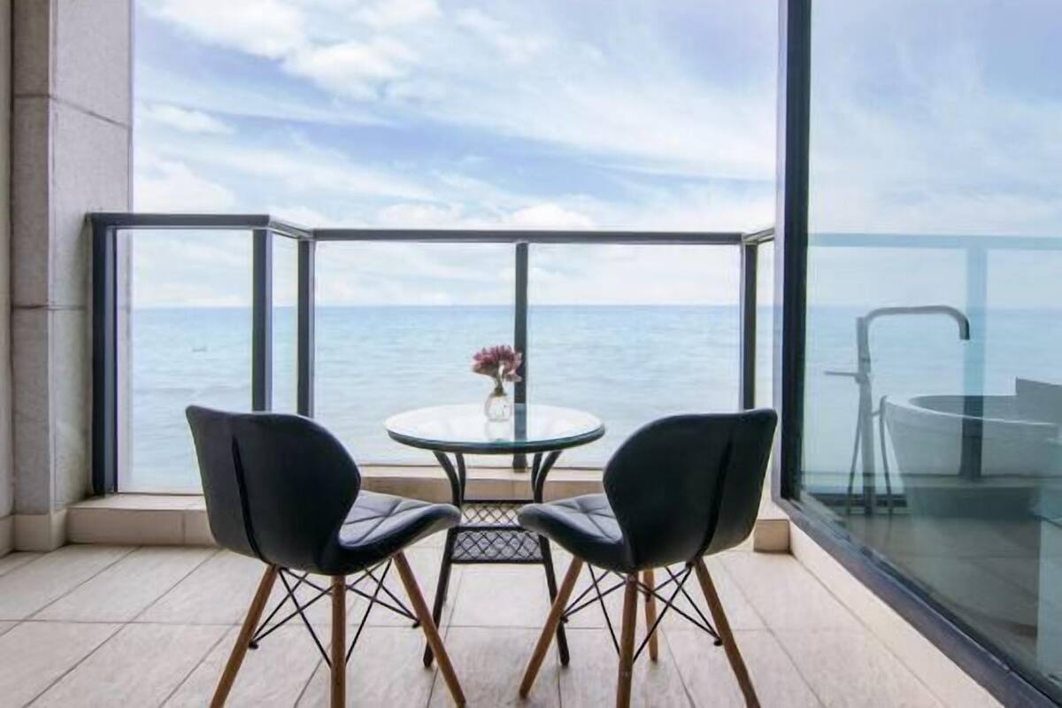 Un balcón con barandilla de vidrio, dos sillas, una mesita redonda con un florero y vista al mar.
