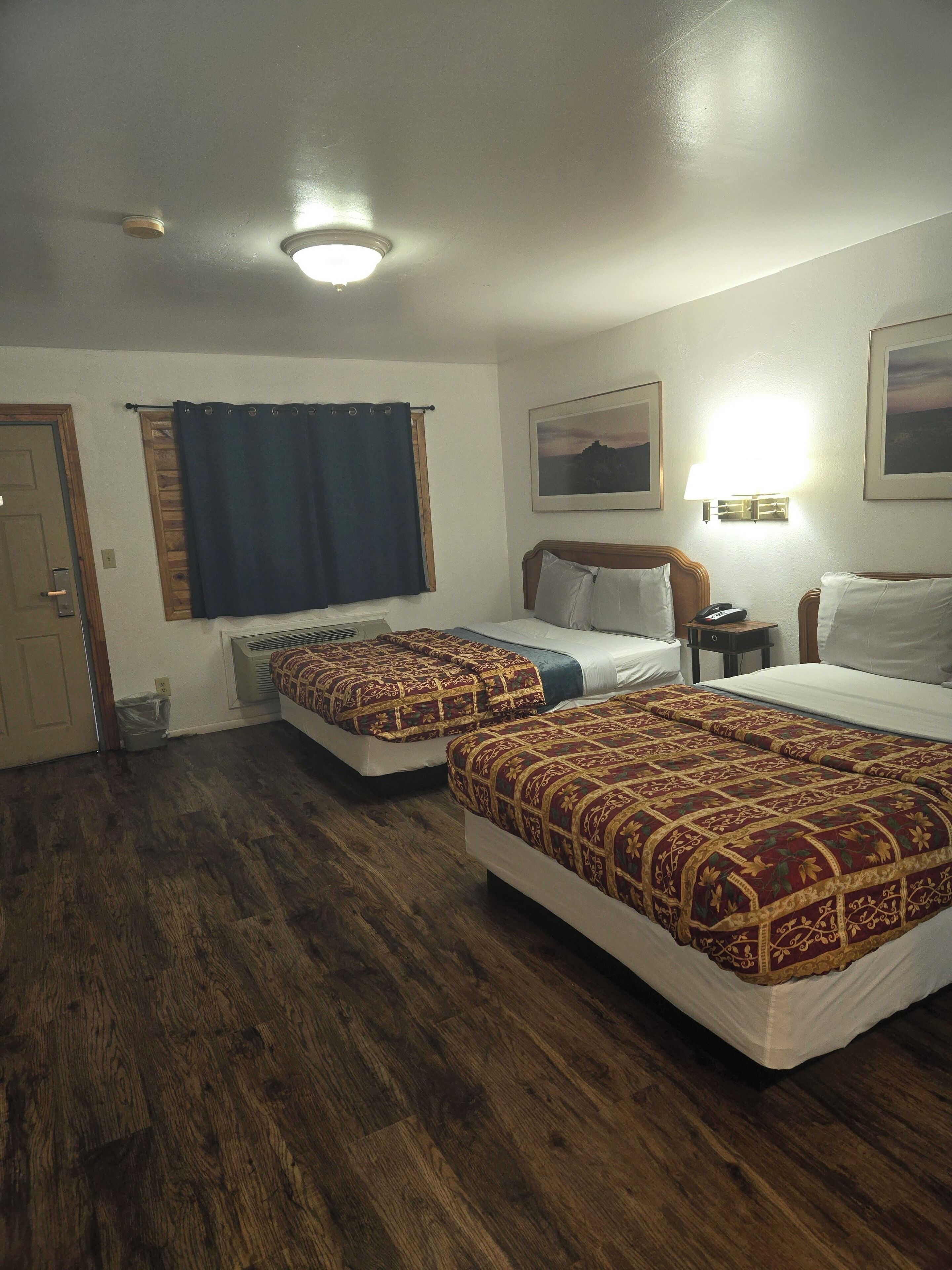 A hotel room with two beds, a wooden floor, a window with a curtain, a wall-mounted lamp, and a door.