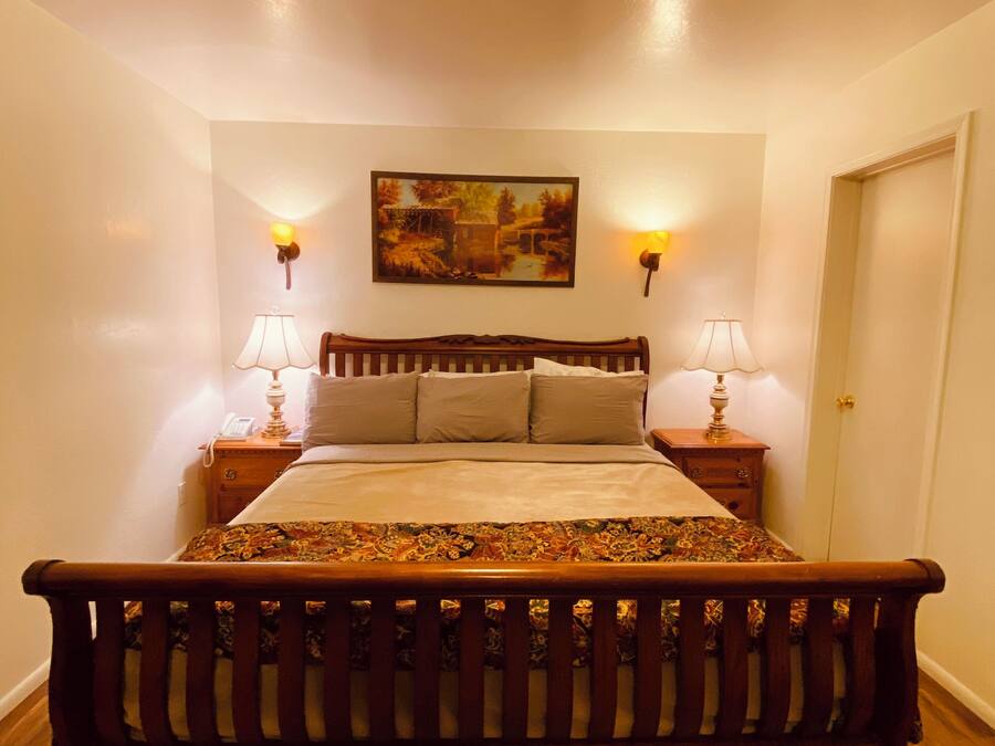 Un dormitorio con cama de madera, dos lámparas de noche, un cuadro en la pared y una puerta a la derecha.