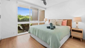 Une chambre avec un grand lit, un ventilateur de plafond, une table de chevet avec une lampe et une vue sur l’extérieur, accessible par une porte-fenêtre coulissante.