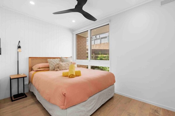 A bedroom with a bed, a bedside table, a ceiling fan, and a window with a view of greenery outside.