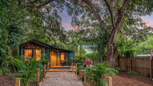 A small, wooden cabin with a porch, surrounded by lush greenery and trees.