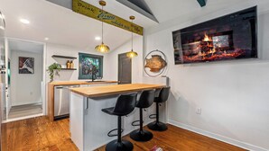 A modern kitchen with a wooden island, black bar stools, pendant lights, and a large TV screen displaying a fireplace.