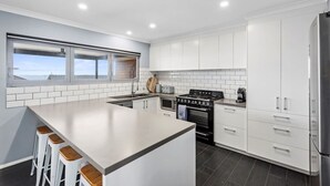 A modern kitchen with white cabinets, a large island with bar stools, a built-in oven, and a view through a window. - Hood Road Beach House- Hamptons inspired sleeps 9 (Indented Head)