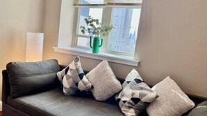 A dark grey sofa with three patterned cushions, a green vase with a plant on a windowsill, and a white wall with a paper on it.