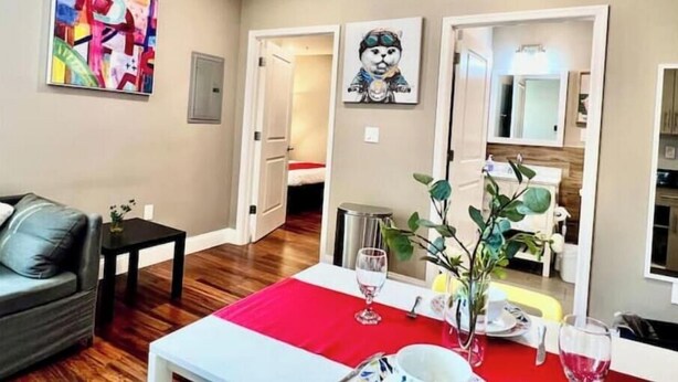 A room with a dining table set for two, a sofa, a small table with a vase, and a painting on the wall.