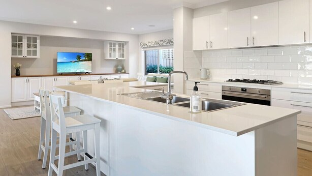 A modern kitchen with white cabinetry, a large island with bar stools, a built-in oven, and a TV displaying a beach scene.