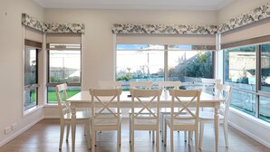 A dining area with a white table and chairs, large windows with a view of a garden and buildings, and a ceiling with recessed lighting.