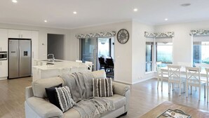 A modern kitchen with white cabinetry, a built-in oven, and a stainless steel refrigerator. There is a dining area with white chairs and a table, and a living room with a beige sofa and patterned throw pillows.