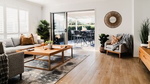 A modern living room with a sofa, coffee table, and a dining area visible through a glass door. - Hamptons Haven; Luxe 4 bedroom, 2 bathroom, WIFI (Armstrong Creek)