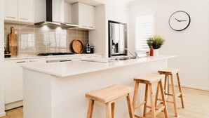 A modern kitchen with white cabinetry, a large island with stools, a stainless steel refrigerator, and a clock on the wall. - Hamptons Haven; Luxe 4 bedroom, 2 bathroom, WIFI (Armstrong Creek)