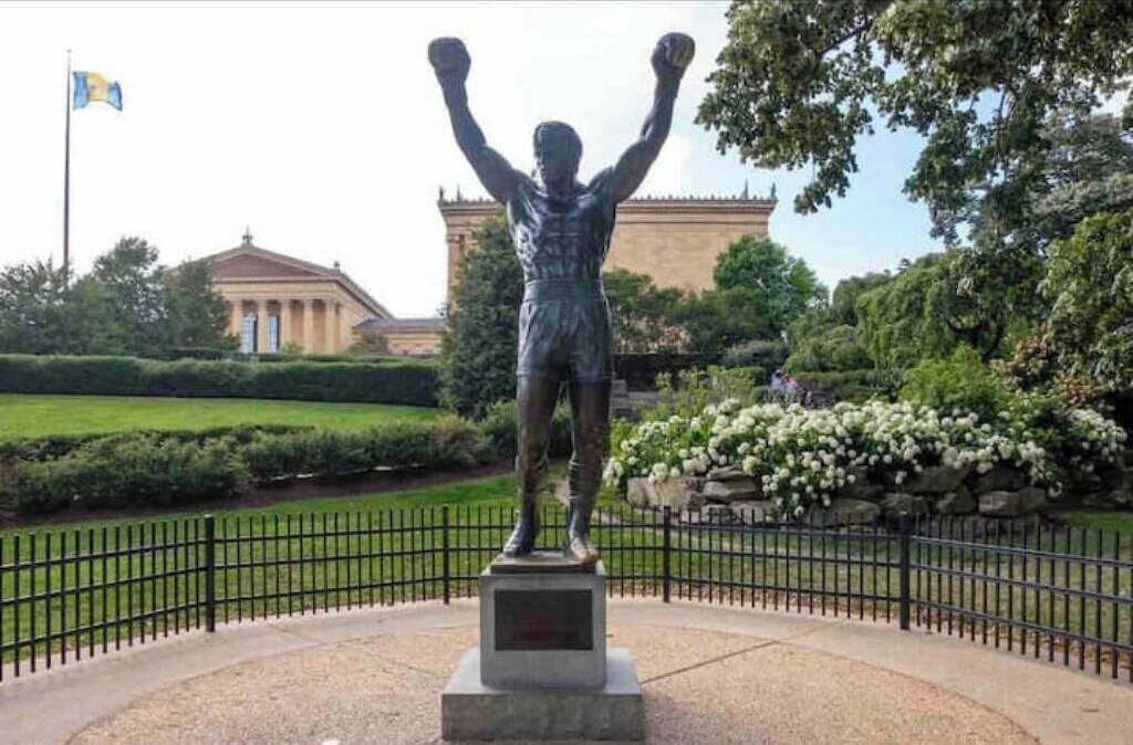 A statue of a boxer with raised arms in a park setting.
