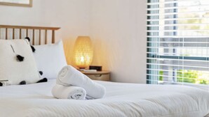 Ein ordentlich gemachtes Bett mit aufgerollten Handtüchern, einer Nachttischlampe und einem Fenster mit Jalousien.