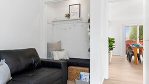 A modern living room with a black leather sofa, a woven rug, a white fireplace mantel with decorative items, and a dining area visible through an open doorway.