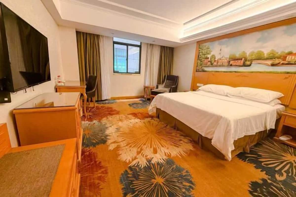 A hotel room with a large bed, a desk with a chair, a television, and a painting on the wall.