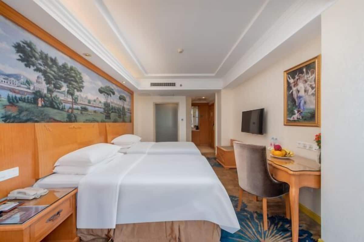A hotel room with two beds, a desk, a chair, a TV, and a painting on the wall.