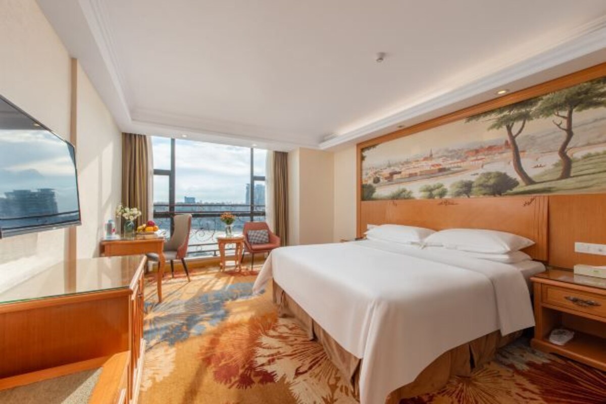 A hotel room with a large bed, a desk, a chair, and a view of the city through large windows.