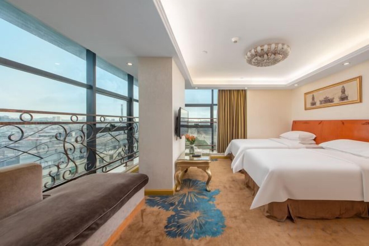 A hotel room with a balcony, two beds, a sofa, a desk with a vase, and a large window.