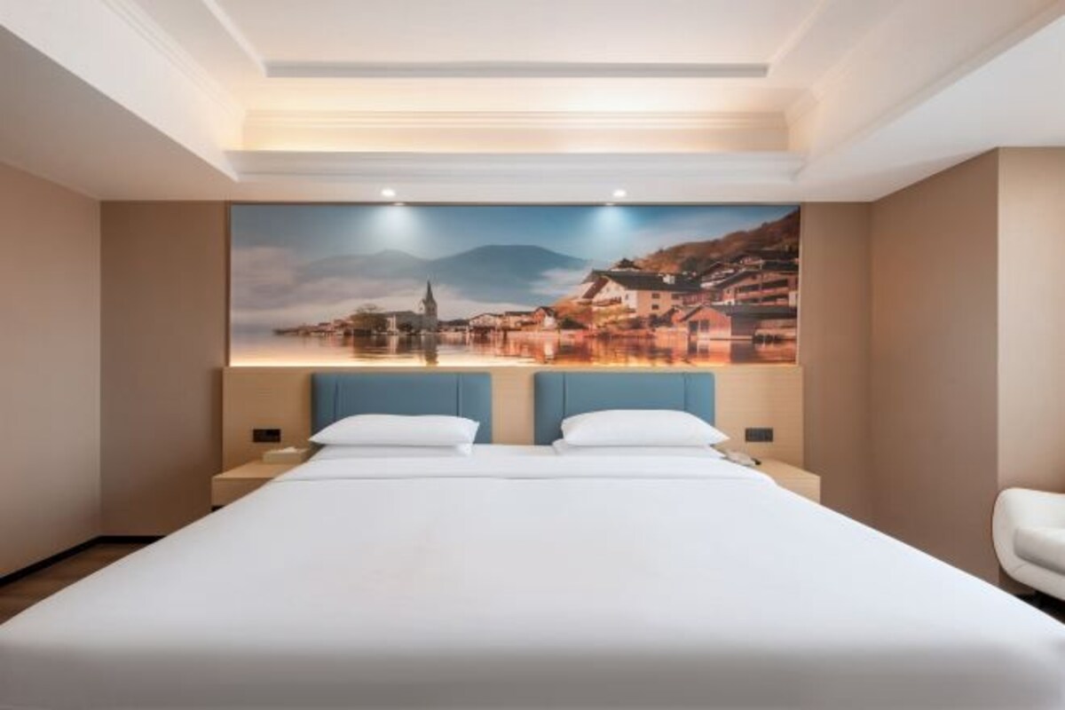 A hotel room with a large bed, a headboard with a cityscape mural, a nightstand, and a white chair.