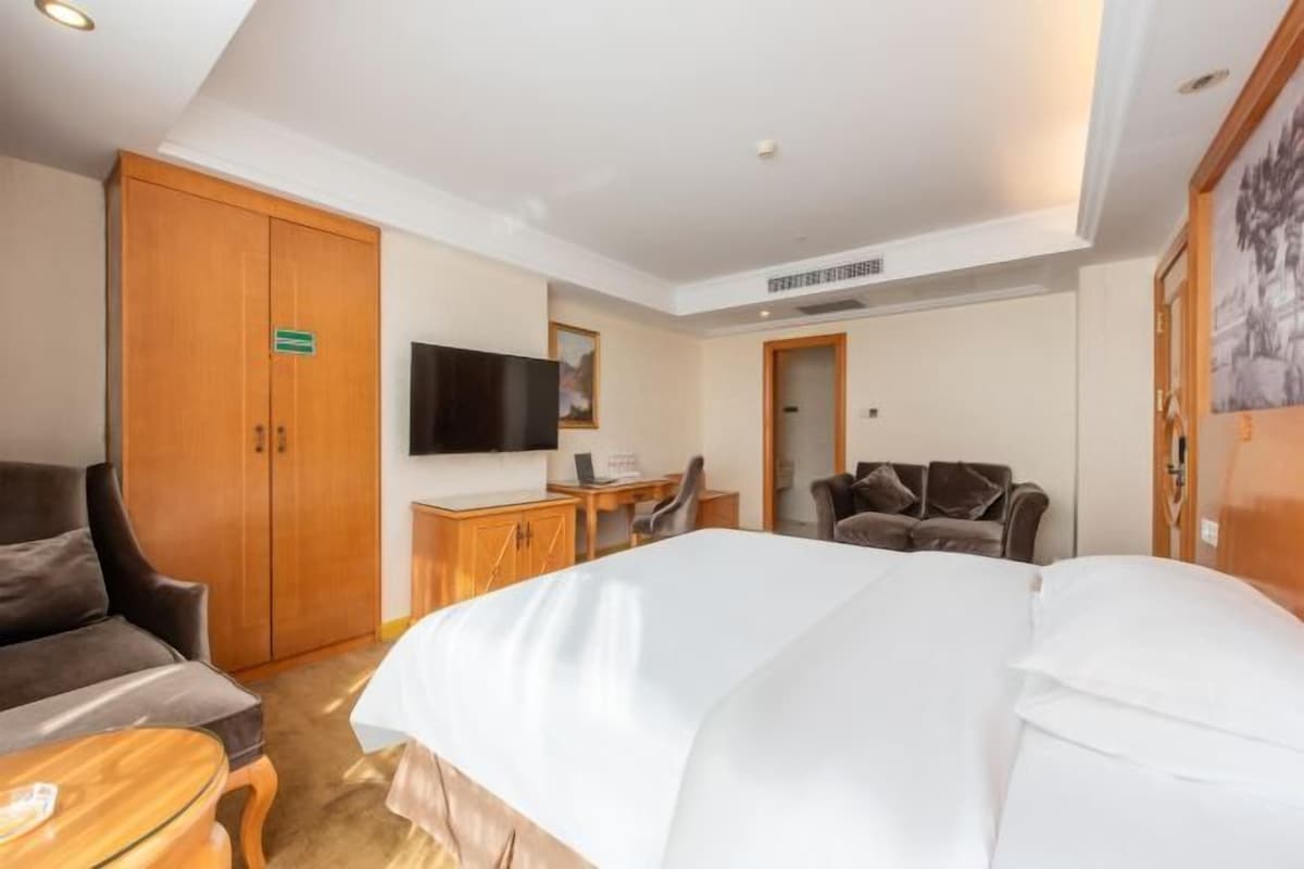 A hotel room with a large bed, a desk with a computer, a TV, a wardrobe, and a sofa.