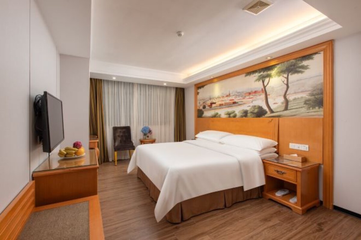 A hotel room with a bed, a TV, a painting on the wall, and a desk with a lamp and a vase of flowers.