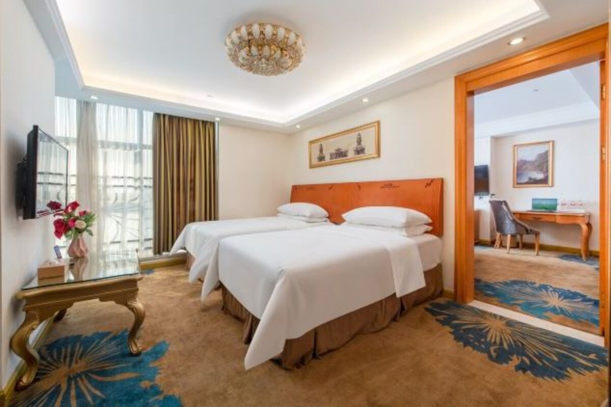 A hotel room with two beds, a desk, a chair, a TV, and a large window with curtains.