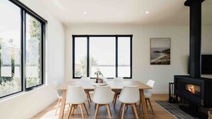 A modern dining area with a large table and white chairs, a black fireplace, and large windows offering a view of the outdoors.