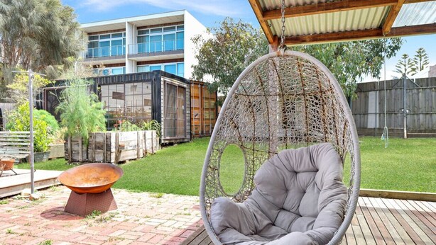 A hanging chair in a backyard with a brick patio, a metal fire pit, and a modern building in the background.