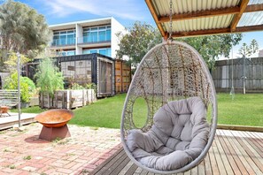 Une chaise suspendue dans un jardin, avec une terrasse en briques, un foyer en métal et un bâtiment moderne en arrière-plan.