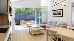 A modern living room with a sofa, coffee table, and chairs, a TV mounted on the wall, and a large sliding glass door leading to an outdoor area. - Beach luxury retreat- 2 bedroom, 2 bathroom, shops (Ocean Grove)