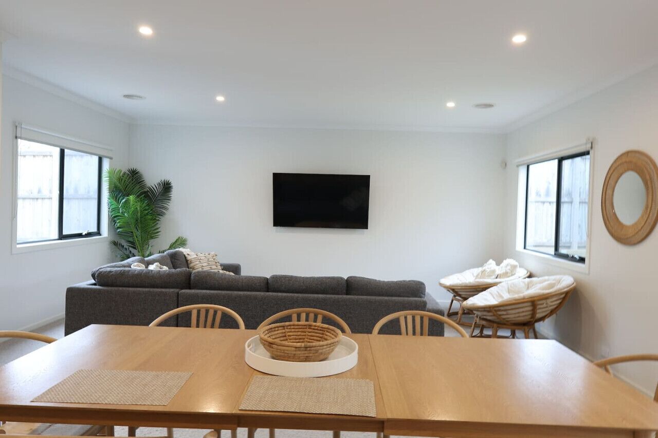 A modern living room with a gray sofa, a dining table with chairs, a large window, and a television mounted on the wall.