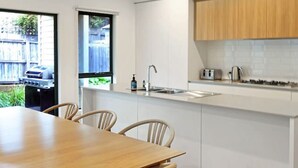 A modern kitchen with white cabinetry, a wooden dining table, and chairs. There is a window with a view of a backyard. - Madeley Family Getaway- Sleeping 8, central, a/c (Ocean Grove)