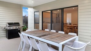 An outdoor patio with a wooden table and white chairs, a grill, and a sliding glass door leading to a kitchen area. - Madeley Family Getaway- Sleeping 8, central, a/c (Ocean Grove)