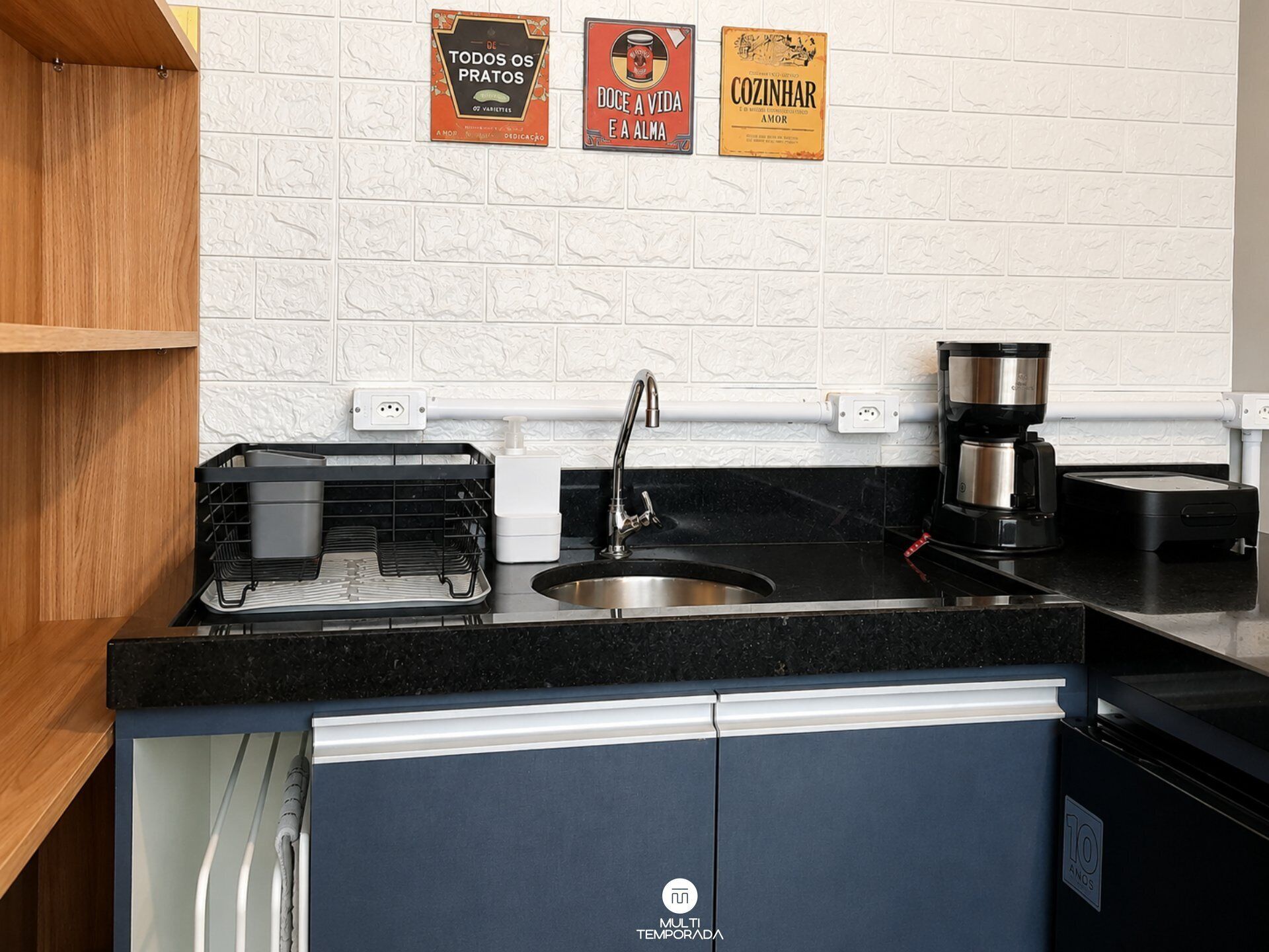 Una cocina con encimera negra, pileta de acero inoxidable y cafetera. Hay tres letreros decorativos en la pared.
