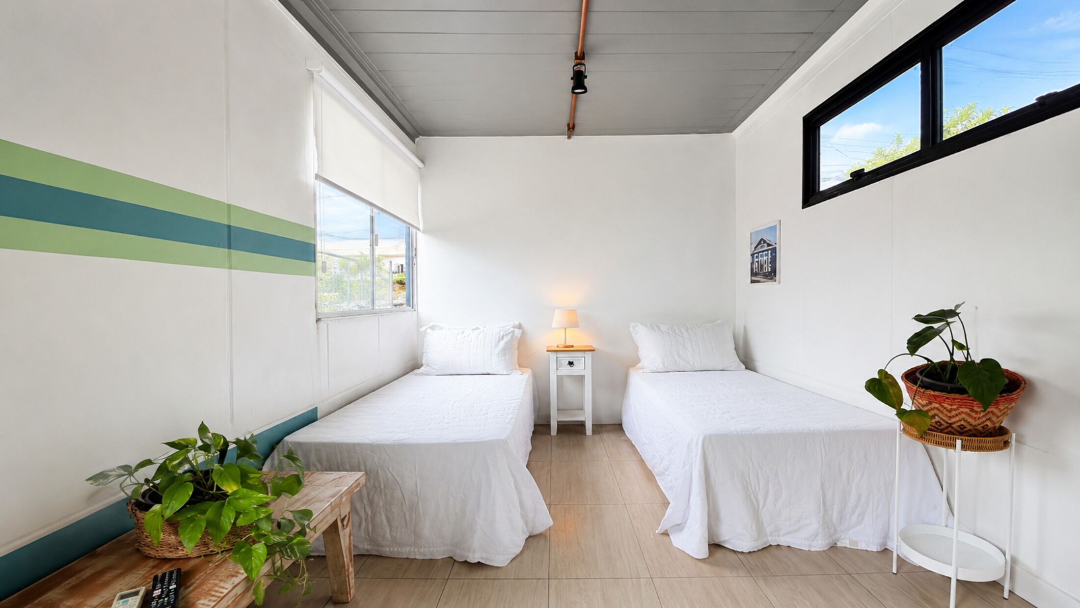 Una habitación pequeña y limpia con dos camas, una mesita de noche, una lámpara, una planta en maceta y una ventana.